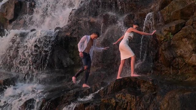 A guy and a girl in a white dress neatly make their way among the rocks under a waterfall during sunset