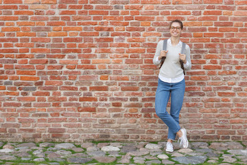 Full-height portrait of young European female standing against red brick wall, leaning to it with...