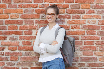 Closeup image of young Caucasian lady with red brick wall behind her, wearing gray backpack, holding arms crossed, looking straight at camera with happy satisfied smile, dressed casually and neutral.