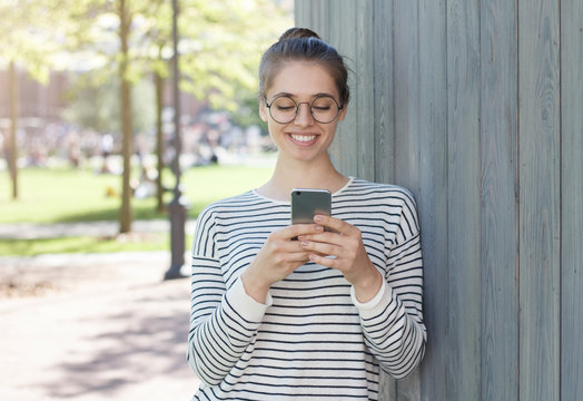 Closeup photo of good-looking Caucasian girl dressed in striped top looking attentively with satisfied smile at display of smartphone, typing, willing to continue friendly joyful communication in net. - Powered by Adobe
