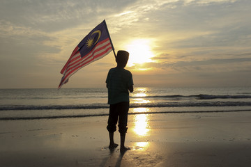 The Concept of Independence Day - a boy holding the Malaysian flag on the shore at sunrise