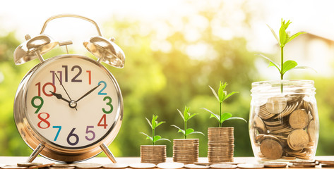 wooden house model and step of coins stacks with tree growing on top and clock alarm, nature background, money, saving and investment or family planning concept, over sun flare silhouette tone.