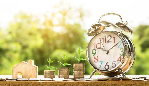 wooden house model and step of coins stacks with tree growing on top and clock alarm, nature background, money, saving and investment or family planning concept, over sun flare silhouette tone.