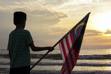 The Concept of Independence Day - a boy holding the Malaysian flag on the shore at sunrise