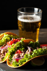 Photo of Mexican tacos with ground meat, beef, beans, onions and salsa on wooden background. Ketchup sauce and lime. A glass o beer in the background.