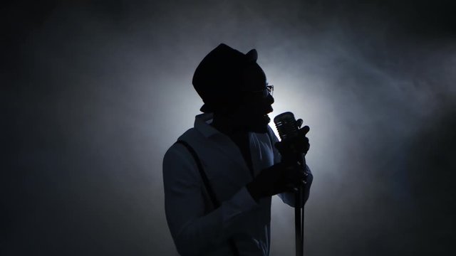 Man african american singer in the smoke and white light sings into a microphone and dance. Black background. Silhouette