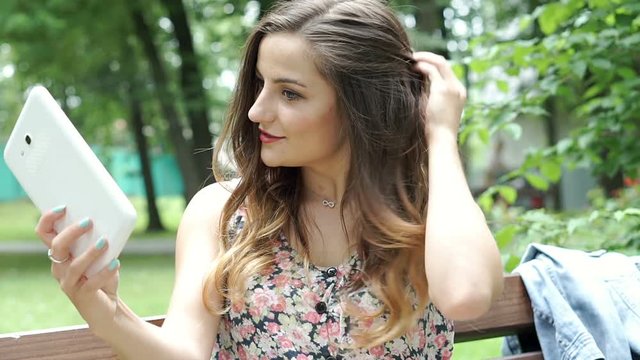 Pretty brunette in floral shirt doing selfies on tablet in the park, steadycam shot
