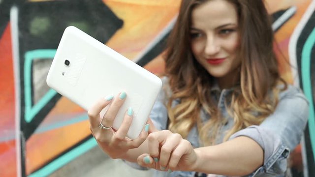 Pretty brunette standing next to the graffiti wall and doing selfies on tablet, steadycam shot
