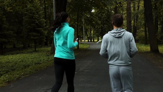 Two young sporty powerful runners synchronous running in autumn Park on asphalt road. They have gadjets: smartphone, iphone, ears, headphones, hands-free headset. Slow motion