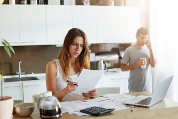 Busy female worker working with pile of documents, reading papers with attentive look, studying diagrams and charts, being absorbed into work while her husband chatting over mobile phone, drinking tea