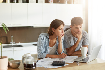 Portrait of family couple looking attentively into screen of laptop, sitting at kitchen table, surrounded with documents, making business reprort, studying diagrams and charts. People, figures, budget
