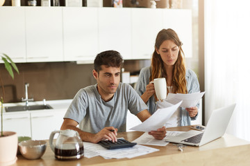 Picture of serious young American couple spending weekend morning in kitchen, doing paperwork, looking through mail and calculating family expenses, paying bills online using laptop computer