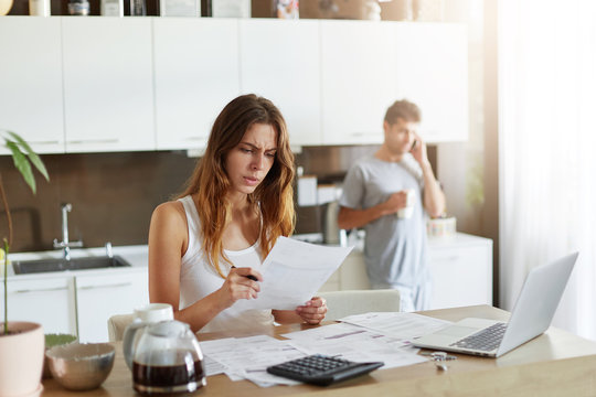 Puzzled female accounter trying to understand what is written in paper, reading it with frustrated look, using calculator and modern laptop computer for making business report and her husband resting