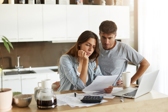 Picture of young wife and husband in kitchen having concentrated looks while reading notification from bank concerning their loan application. Caucasian couple calculating expenses in the morning