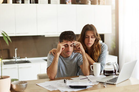 People, relationships, debts, financial stress and economic crisis. Unemployed young Caucasian man feeling depressed, sitting at kitchen table over unpaid bills while his wife trying to cheer him up