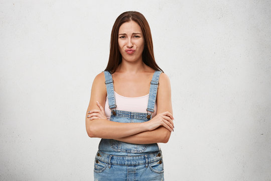 Picture of attractive young Caucasian female with dark straight hair posing in studio, keeping arms folded, having offended look, pouting lips, showing her dislike, negative attitude and rigidity