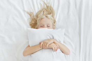 Carefree restful little girl lying on white bedclothes, embracing pillow while having pleasant...