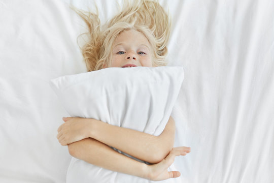 Funny little girl with blonde hair and blue eyes, having fun in bed, embracing white pillow, going to fall asleep. Happy small child with pillow at home, relaxing at bedroom. Children lifestyle
