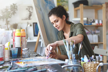 Picture of serious concentrated young Caucasian female artist sitting at desk with painting accessories, holding tube of oil paint, mixing colors on palette; unfinished painting on canvas near her - obrazy, fototapety, plakaty