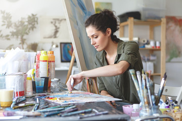 Serious brunette young beautiful woman sitting in art studio, taking colorful paints from tube...