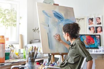 Art, creativity, hobby, job and creative occupation concept. Rear view of busy female artist sitting on chair in front of easel, painting with fingers, using white and blue oil or acrylic paint - obrazy, fototapety, plakaty