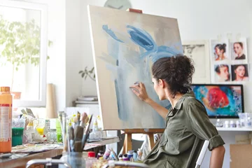 Fotobehang Art, creativity, hobby, job and creative occupation concept. Rear view of busy female artist sitting on chair in front of easel, painting with fingers, using white and blue oil or acrylic paint © Wayhome Studio