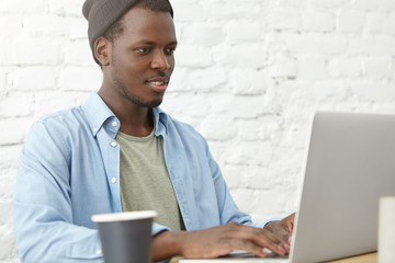 Portrait of black male in stylish clothes using free internet connection at cafeteria, working at laptop, surfing social networks and drinking coffee. Businessman working with modern device at cafe
