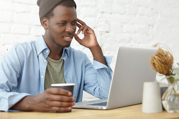 Picture of fashionable young African American male programmer, blogger, freelancer or startupper using laptop computer at coworking space for remote work and drinking coffee, having happy look