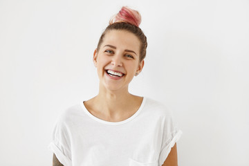Sincere joy, happiness, youth and carefree life. Indoor shot of lovely hipster girl with hair bun dyed pink having fun, laughing out loud at some joke or funny story, dressed in stylish white t-shirt
