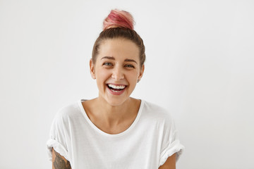 People, youth, joy, fun, happiness and relaxation concept. Indoor portrait of trendy-looking hipster girl with hair knot dyed pink smiling cheerfuly, can't help but laughing while watching good comedy