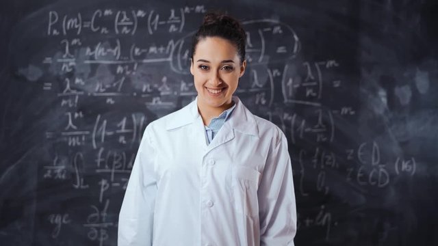 Young student female in white robe solve hard and heavy to understand math equation on black chalk board, She turn to camera and smile. Smart, clever, intelligent scientist woman concept.