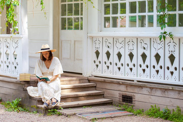 woman in a white dress and a straw hat reading a book on the porch of a rural house