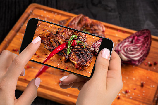 Photographing food concept - tourist takes picture of ready-to-eat beef steak dish on cutting wooden board on smartphone.