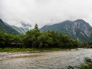 Naklejka na ściany i meble The view of the stream flows down through the forest on the mountain with cloud background at Kamikochi Japan