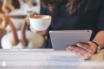 Closeup image of a woman holding and using tablet pc while drinking coffee in modern cafe