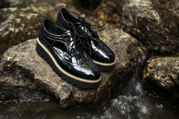 Black shiny patent leather women's brogues shoes on old gray stones, near river in a forest or park. Fashion advertising shoes photos.