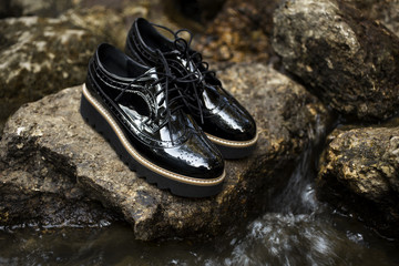 Black shiny patent leather women's brogues shoes on old gray stones, near river in a forest or...