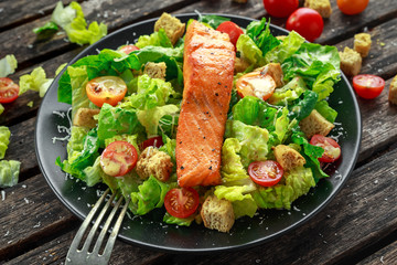 Caesar Salmon Salad with croutons, parmesan cheese, tomatoes, dressing and pepper in a black plate on wooden table.