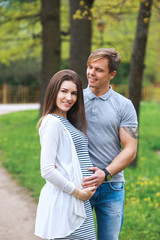 Handsome man and his beautiful pregnant wife are hugging and smiling while standing in park