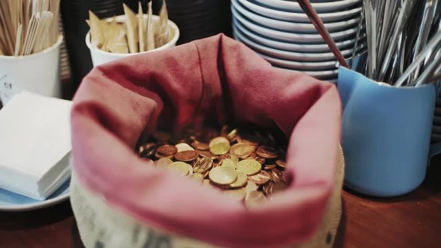 Hand held selective focus shot of camera approaching bag of coins or change, top of bar or cafeteria counter, tips for barista or waiter, with plates and cuttlery, concept economy or service industry
