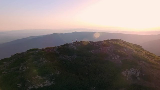 Beautiful summertime lifestyle drone video of attractive young hipster millennial couple descending mountain ridge, with purple pink sunset or sunrise bursting in colors behind them