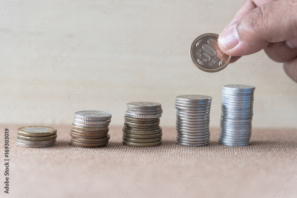 Wall mural Saving money concept, Hand hold coin, roll stair step of coins on white table with gray wood light background.