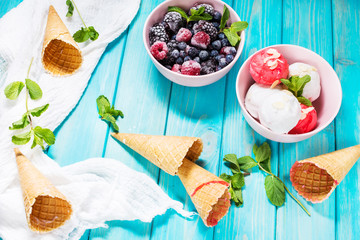 Ice cream in bowl, berries and waffle cones on blue wooden table.
