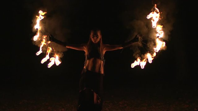 Young girl dances with burning torches made in shape of fans at fire show.