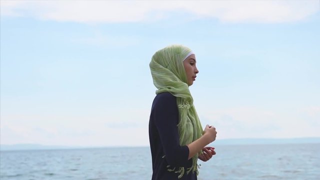 A young Muslim woman in a scarf that covers her hair doing exercises near the sea, a lady runs in sport sneakers over small stones that are near the water