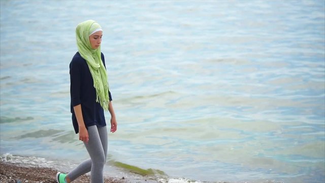 A young Muslim woman in a veil, which covers her hair, walks along the shore along the sea, the lady looks thoughtfully into the distance, she looks serious and focused