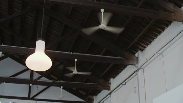 Couple of ventilators attached to wooden and metal ceiling of an industrial building spin and turn swiftly in air to creat current of wind, with white modern lamp swinging nearby