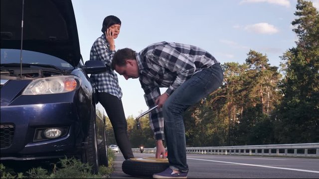 handsome caucasian man changing a tire on the side of the road. Young woman wearing casual clothes has phone conversation on smartphone . Female looking on the guy and smiling.