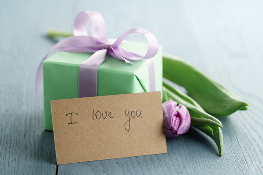 green gift box with purple bow and tulip on blue wood background with i love you greeting card