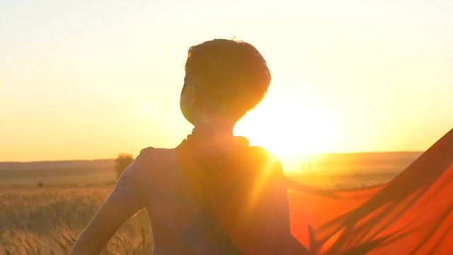 A boy with a cape of a superman stands in the golden fields of wheat looking at the horizon